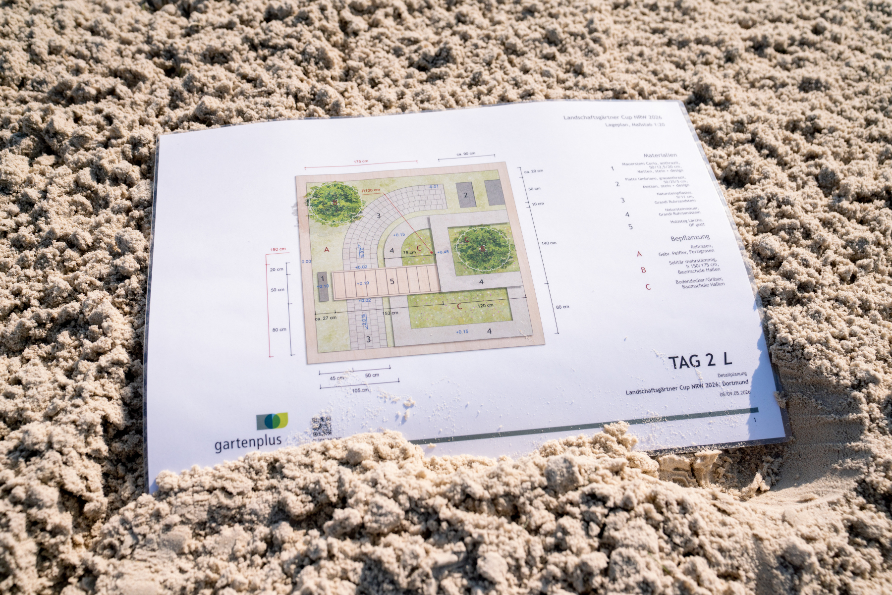 Landschaftsgärtner Cup NRW 2026, Detailplanung auf Sand mit gartenplus Im Sand liegt ein ausgedruckter Gartenplan zum Landschaftsgärtner Cup NRW 2026 mit gartenplus-Logo und der Beschriftung TAG 2 L.