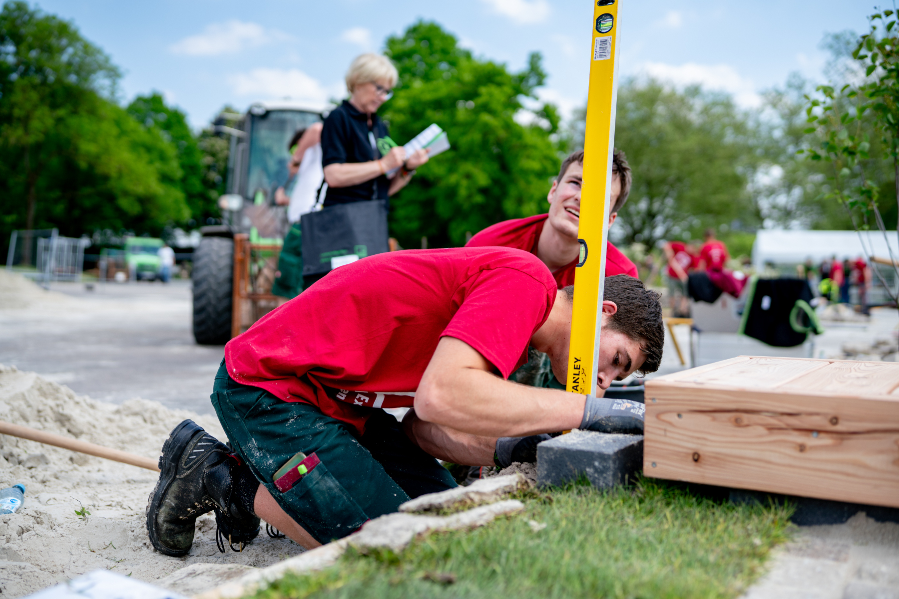 Gartenbau mit Wasserwaage und Holzrahmen, junge Männer bei Vermessungsarbeit Zwei junge Männer in roten Shirts richten mit einer gelben Wasserwaage einen Holzrahmen im Garten aus, während im Hintergrund weitere Helfer arbeiten.