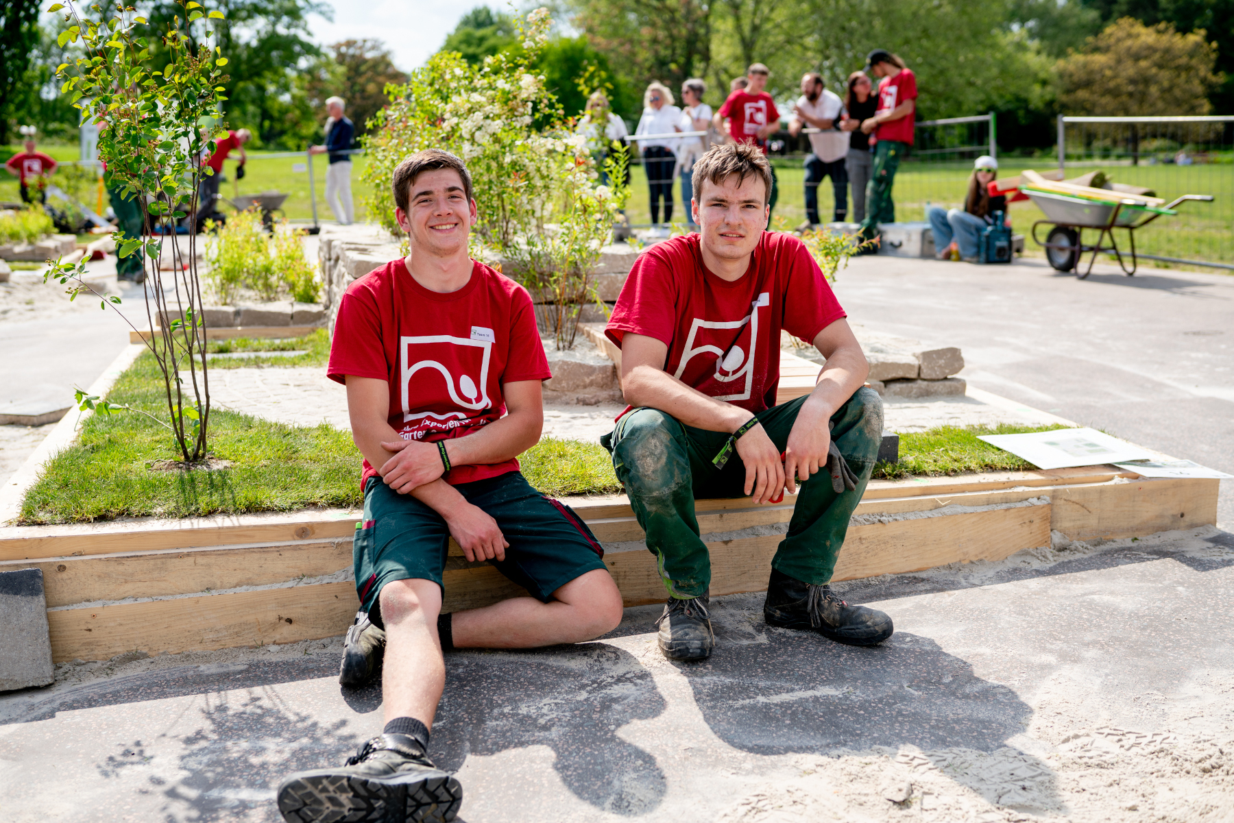 Gartenbau-Team in roten Shirts am neu angelegten Beet, junge Helfer im Freien Zwei junge Helfer in roten Shirts sitzen am Rand eines neu angelegten Beets, während im Hintergrund weitere Personen an der Gartenanlage arbeiten.