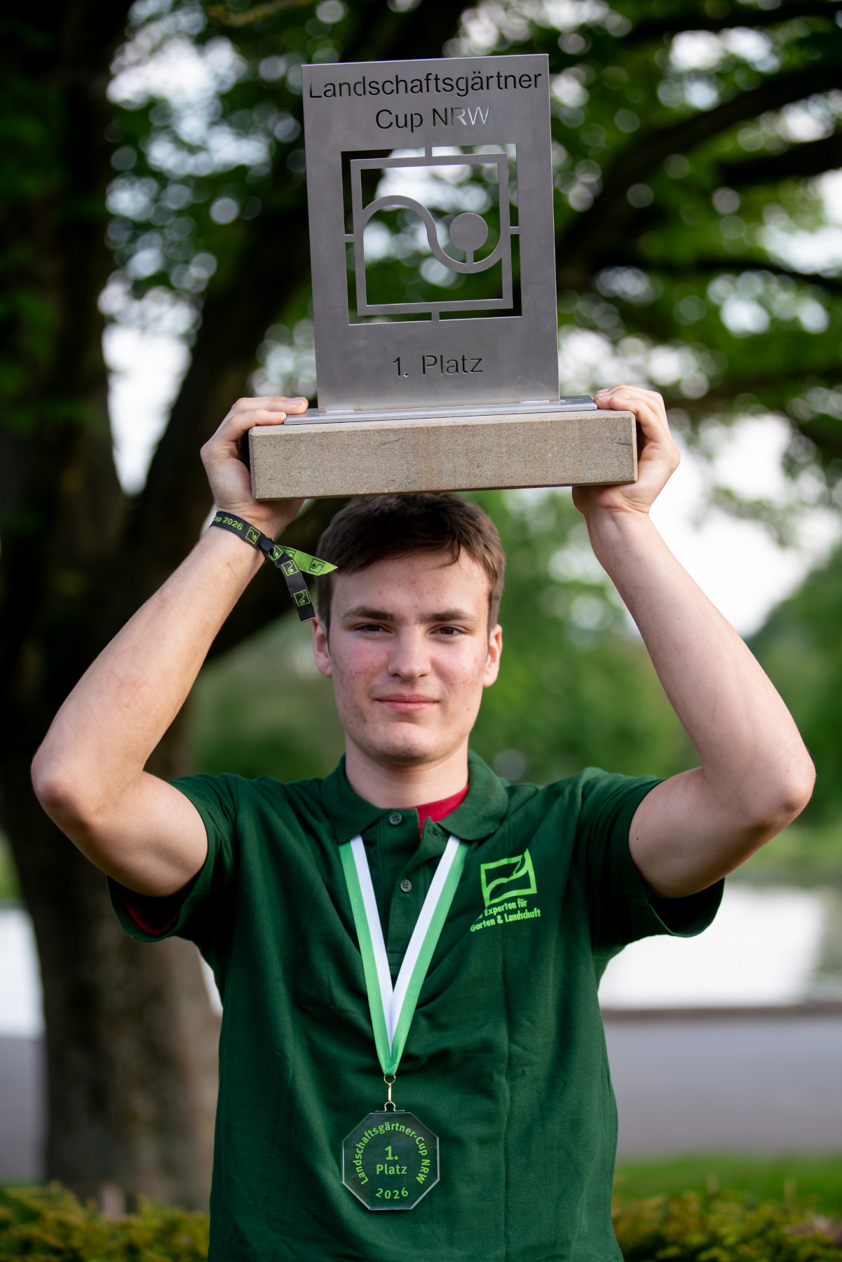 Landschaftsgärtner Cup NRW, Sieger mit Pokal und 1. Platz-Auszeichnung Ein junger Mann hält beim Landschaftsgärtner Cup NRW den Pokal für den 1. Platz über dem Kopf, mit Medaille und grünem Polohemd.