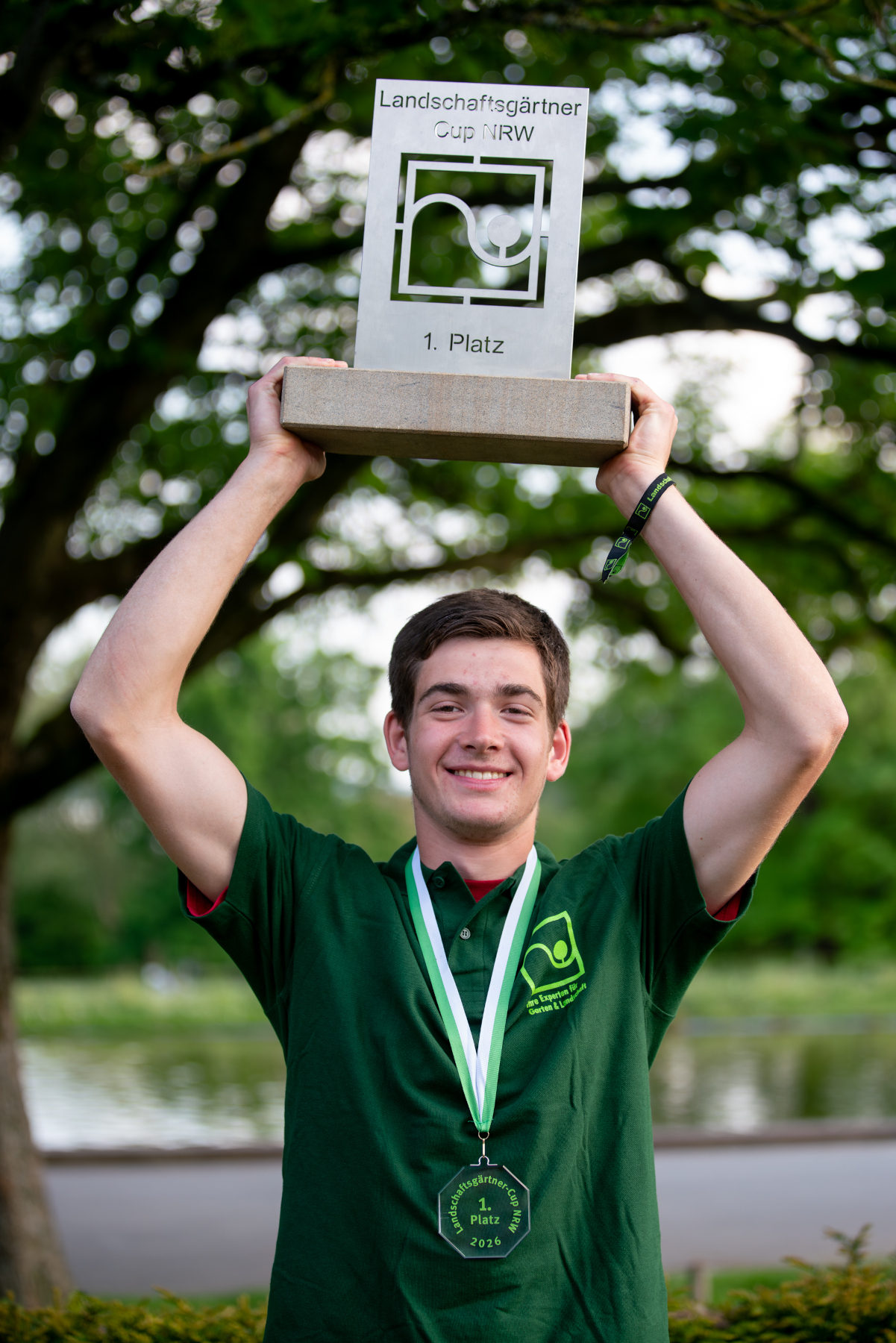 Landschaftsgärtner Cup NRW, Sieger mit Pokal und Medaille im Grünen Ein junger Mann hält den Pokal des Landschaftsgärtner Cup NRW mit der Aufschrift 1. Platz hoch und trägt Medaille sowie grünes Shirt im Freien.