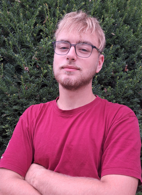 Ein lächelnder junger Mann im roten Poloshirt steht an einem windigen Hang mit hohem Gras unter klarem blauem Himmel und blickt in die Kamera.